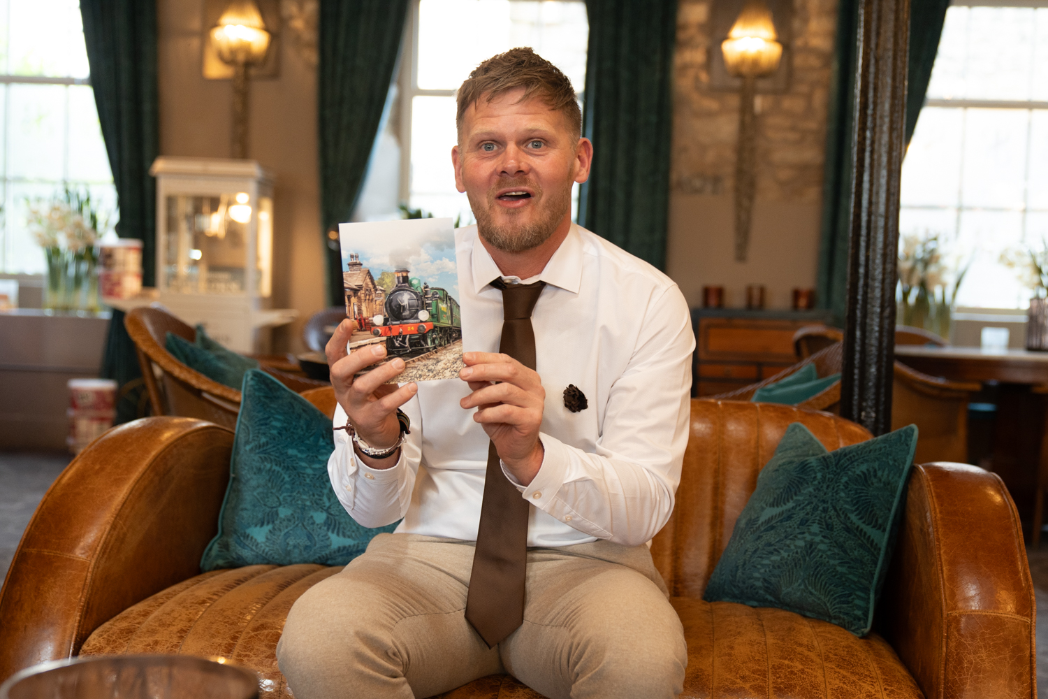 The groom sits on a leather sofa in a hotel lounge, grinning as he reads a card — completely delighted, tie on, boutonnière pinned